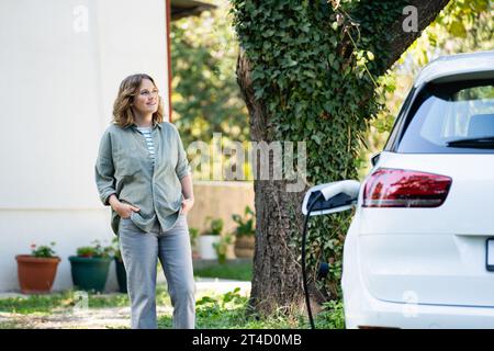 Frau neben einem Elektroauto im Hof eines Landhauses. Stockfoto