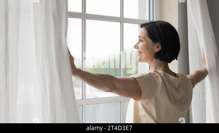 Seitliche Ansicht, ausgeruhte Frau mittleren Alters, die Vorhänge öffnet Stockfoto