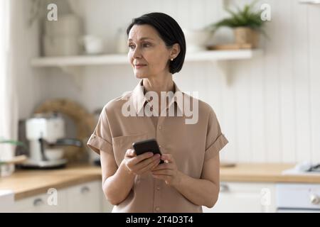 Nachdenkliche Frau mittleren Alters, die mit Smartphone in der Küche steht Stockfoto