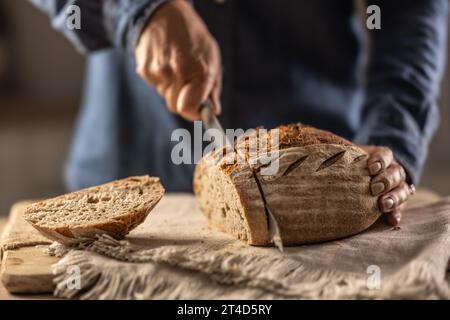 Frau, die frisches Brot auf der Theke in der Küche schneidet - Nahaufnahme. Der mittlere Teil des Weibchens ist mit Messer und gebackenem Brot. Stockfoto