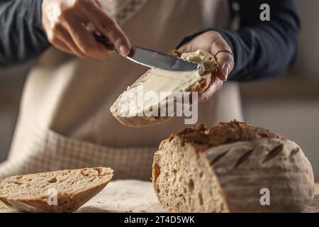 Eine Frau macht köstliches Brot, verteilt Frischkäse mit einem Besteckmesser - Nahaufnahme. Frau Hände, die Frischkäse auf Brotscheibe verteilen. Stockfoto