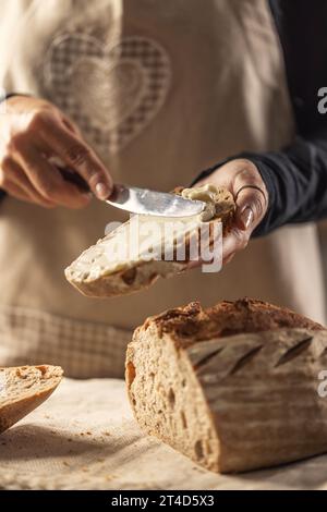 Eine Frau macht köstliches Brot, verteilt Frischkäse mit einem Besteckmesser - Nahaufnahme. Frau Hände, die Frischkäse auf Brotscheibe verteilen. Stockfoto