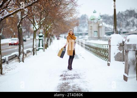 Glückliche moderne Frau in braunem Hut und Schal in Kamelmantel mit Handschuhen und Einkaufstaschen draußen in der Stadt im Winter. Stockfoto