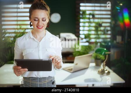 Umweltfreundliches Immobiliengeschäft. Lächelnde 40-jährige Maklerin im modernen grünen Büro in weißer Bluse mit digitalem Tablet. Stockfoto