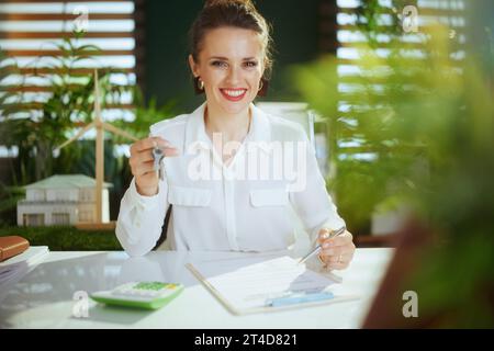 Es ist Zeit, weiterzumachen. Glückliche, moderne 40-jährige Geschäftsfrau im modernen grünen Büro in weißer Bluse mit Taschenrechner, Zwischenablage, Dokument und Schlüsseln. Stockfoto