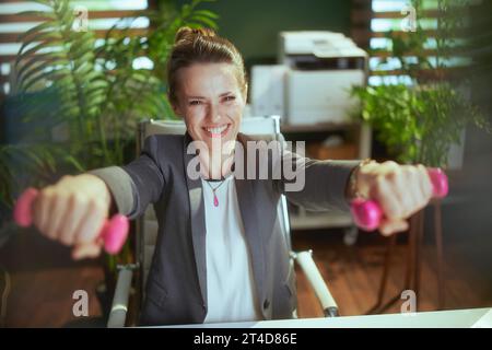 Nachhaltiger Arbeitsplatz. Lächelnde, moderne Geschäftsinhaberin bei der Arbeit mit Kurzhanteln. Stockfoto