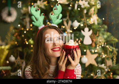 Glückliche elegante 40-jährige Frau mit festlichem heißem Schokoladencocktail und lustigem Rentiergeweih Kopfschmuck im traditionellen Weihnachtspullover nahe C Stockfoto