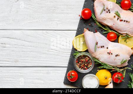 Rohe Hähnchenschenkel mit Tomaten, Kirsch, Zitrone, Knoblauch, Gewürzen und Kräutern auf schwarzem Schieferbrett auf weißem hölzernem Hintergrund. Draufsicht mit Kopierbereich Stockfoto