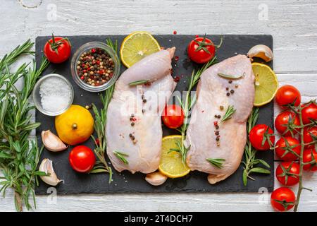 Rohe Hähnchenschenkel mit Tomaten, Kirsch, Zitrone, Knoblauch, Gewürzen und Kräutern auf schwarzem Schieferbrett auf weißem hölzernem Hintergrund. Draufsicht. Kochen von Fleisch für Stockfoto