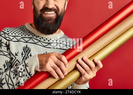 Beschnittener glücklicher bärtiger Mann im Winterpullover, der Geschenkpapier auf rotem Hintergrund hält, Frohe Weihnachten Stockfoto