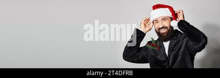 Porträt eines fröhlichen, gutaussehenden Mannes im Anzug mit weihnachtsmütze auf grauem Hintergrund, Weihnachtsbanner Stockfoto