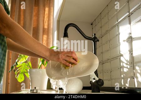 Die Hände einer Frau waschen zu Hause das Geschirr. Teller unter einem Wasserstrahl im Sonnenlicht. Heimroutine. Stockfoto