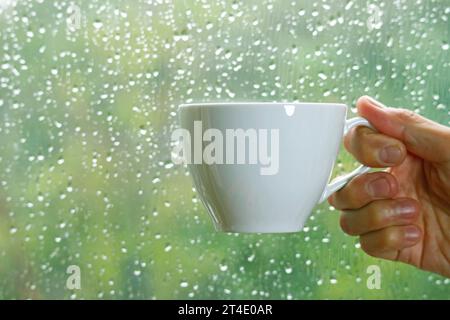 Hand hält eine Tasse Kaffee auf dem Glas des Fensters mit Regentropfen im Hintergrund Stockfoto