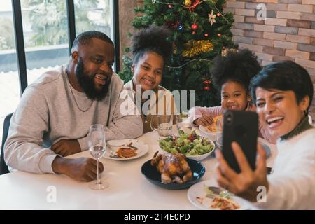 Fröhliche afroamerikanische Familie genießt weihnachten und das Mittagessen im neuen Jahr, während die Frau Selfie auf dem Smartphone mit Familienmitgliedern zu Hause gegen geschmückten Weihnachtsbaum nimmt Stockfoto