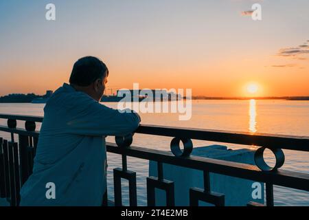 Grauhaariger älterer Mann bewundert den Sonnenuntergang am Ufer des Flusses. Die Einsamkeit der Alten. Sehnsucht und Liebe zum Reisen, Traum von einer Kreuzfahrt. Stockfoto