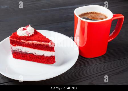 Ein Stück roter Samtkuchen mit cremeweißer Zuckerguss auf einem Holztisch, begleitet von einer dampfenden Tasse aromatischem Kaffee. Stockfoto