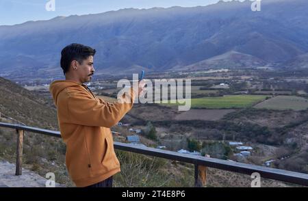 Brauner latino-Mann in einem senffarbenen Kapuzenpullover, der mit seinem Handy ein Foto von einer unglaublichen Landschaft aus Tal und Bergen macht Stockfoto