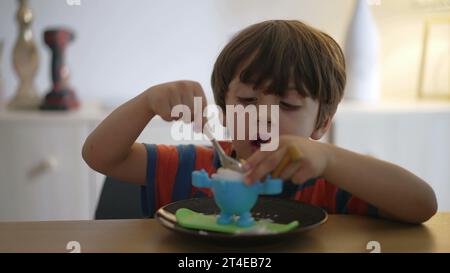 Ein kleiner Junge isst weich gekochtes Ei, oeuf a la coque. Das Kind isst eine leichte Mahlzeit allein mit einem Löffel Stockfoto