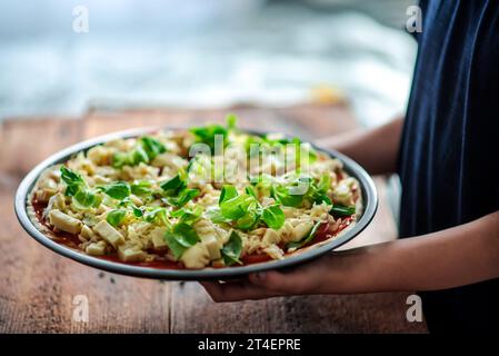 Rohe Pizza in runder Form mit viel Käse und Kräutern in den Händen eines Kindes. Stockfoto