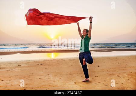 19.02.2019 Goa, Indien. Yogalehrer steht in Asana (yog) auf einem Bein und hält einen flatternden Schal am Strand von Goa Stockfoto