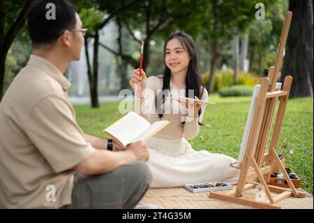 Ein fröhliches und liebenswertes asiatisches Mädchen mag es, mit ihrem Vater in einem grünen Park zu malen, zu picknicken und eine glückliche Zeit miteinander zu verbringen. Stockfoto