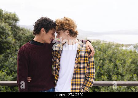 Glückliche, abwechslungsreiche, schwule männliche Paare, die sich auf dem Balkon mit Blick auf den Wald, Kopierraum umarmen Stockfoto