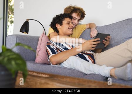 Glückliches, abwechslungsreiches, schwules Paar, das ein Tablet benutzt und zu Hause umgeht Stockfoto