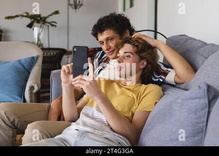 Glückliches, vielseitiges schwules Paar, das Smartphone nutzt und zu Hause umgeht Stockfoto