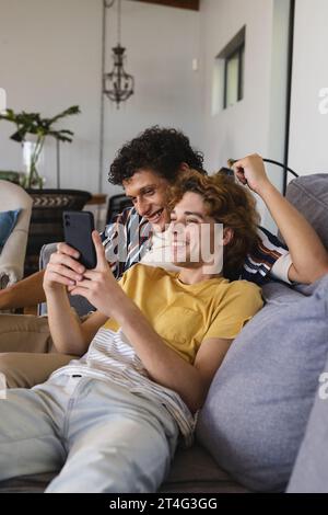 Glückliches, vielseitiges schwules Paar, das Smartphone nutzt und zu Hause umgeht Stockfoto