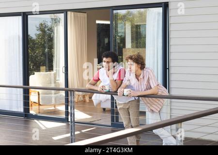 Glückliches, abwechslungsreiches, schwules Paar, das Kaffeetassen auf dem Balkon hält Stockfoto