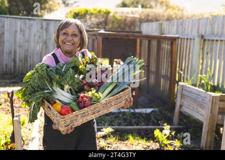 Porträt einer glücklichen älteren birassischen Frau, die einen Korb mit Gemüse im sonnigen Garten hält Stockfoto