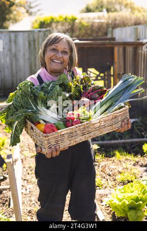 Porträt einer glücklichen älteren birassischen Frau, die einen Korb mit Gemüse im sonnigen Garten hält Stockfoto