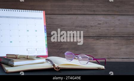 Schreibtischkalender mit Notizblöcken, Stift und Brille auf dem Schreibtisch, Hintergrund aus Holz. Stockfoto