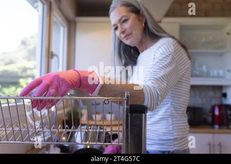 Glückliche ältere kaukasische Frau, die zu Hause Geschirr in der Küche wäscht Stockfoto