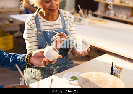 Glückliche zweirassige ältere Töpferin mit grauen Haaren, glasierenden Tonkännchen und lächelndem Töpferstudio Stockfoto