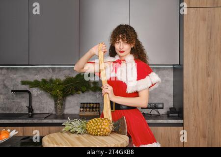 Eine junge Frau in weihnachtlichem rotem Kleid, die sich in der Küche auf eine Axt stützt Stockfoto