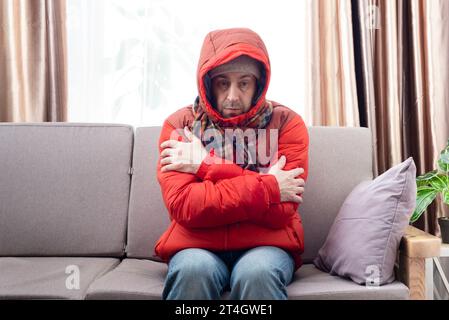 Männliches Model mittleren Alters in rotem Parka und kariertem Schal, der kalt wird und auf dem Sofa im kalten Wohnzimmer sitzt Stockfoto