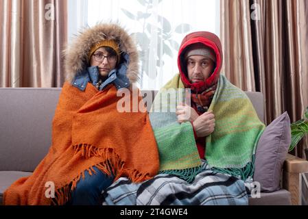 Trauriges Paar mittleren Alters in warmen Kleidern, das auf dem Sofa in ihrem Wohnzimmer kalt wird Stockfoto