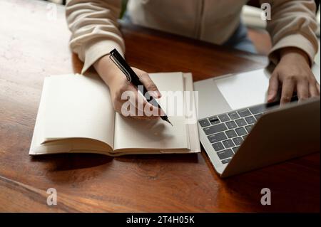 Zugeschnittenes Bild einer Frau, die an ihrem Laptop arbeitet und Notizen in ihrem Notizbuch am Schreibtisch macht. Konzept des Arbeitsbereichs Stockfoto