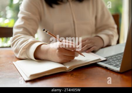 Das Nahbild einer asiatischen Studentin in lässiger Kleidung macht Hausaufgaben, arbeitet an ihrem Laptop und macht Notizen in ihrem Buch an einem Tisch Stockfoto