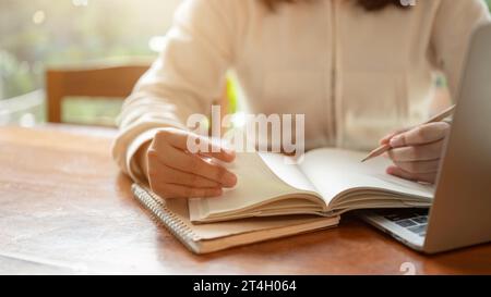 Zugeschnittenes Bild einer Frau, die einen Bleistift hält, in einem Buch an einem Holztisch schreibt. Tagebuch führen, Hausaufgaben machen, Listen erstellen, Online-Unterricht halten Stockfoto