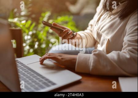 Zugeschnittenes Bild einer asiatischen Frau, die ihr Smartphone hält, während sie auf ihrer Laptop-Tastatur schreibt und drinnen an einem Tisch arbeitet. Menschen- und Technologiekonzepte Stockfoto