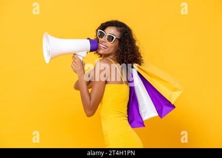 Schöne Frau, die Einkaufstaschen trägt und auf Megaphon im isolierten gelben Farb-Studiohintergrund für Sommerverkauf spricht Stockfoto