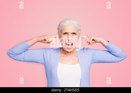 Schöne wütende grauhaarige alte Dame, die lässig ihre Ohren mit Fingern schließt und brüllt. Isoliert über pastellviolettem Hintergrund Stockfoto