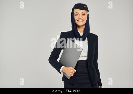 Positive Stewardess der arabischen Fluggesellschaften in Uniform mit einem Laptop, der vor grauer Kulisse in die Kamera lächelt Stockfoto
