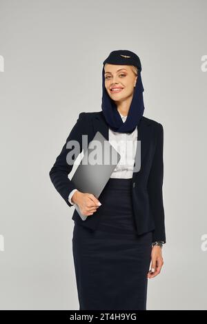 Charmante und elegante Stewardess der arabischen Fluggesellschaft hält einen Laptop und lächelt in die Kamera auf Grau Stockfoto