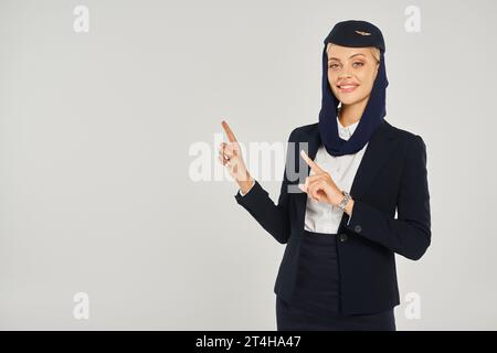 Charmante Stewardess in arabischen Fluggesellschaften lächelt in die Kamera und zeigt mit den Fingern auf Grau Stockfoto
