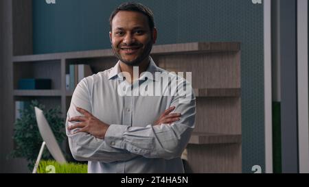 Geschäftsporträt im Büro erwachsener indischer Mann amerikanischer Mann ethnischer Geschäftsmann Unternehmer Arbeitgeber mit überkreuzten Händen posiert am Arbeitsplatz modern Stockfoto