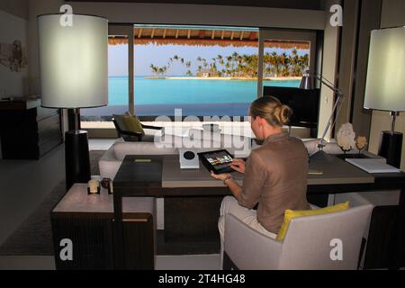 Seychellen, La Digue - 05. Oktober 2022: Frau, die ein Tablet in einem Hotelzimmer benutzt, mit dem Meer aus dem Fenster Stockfoto