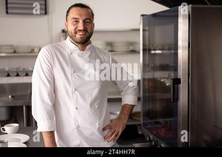 Porträt eines lächelnden kaukasischen Küchenchefs mit Hand auf Hüfte, der in der Küche in der Kochschule steht Stockfoto